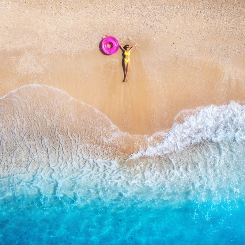 Vista aérea de la hermosa joven acostada con anillo de natación rosa en la playa de arena cerca del mar con olas al atardecer.