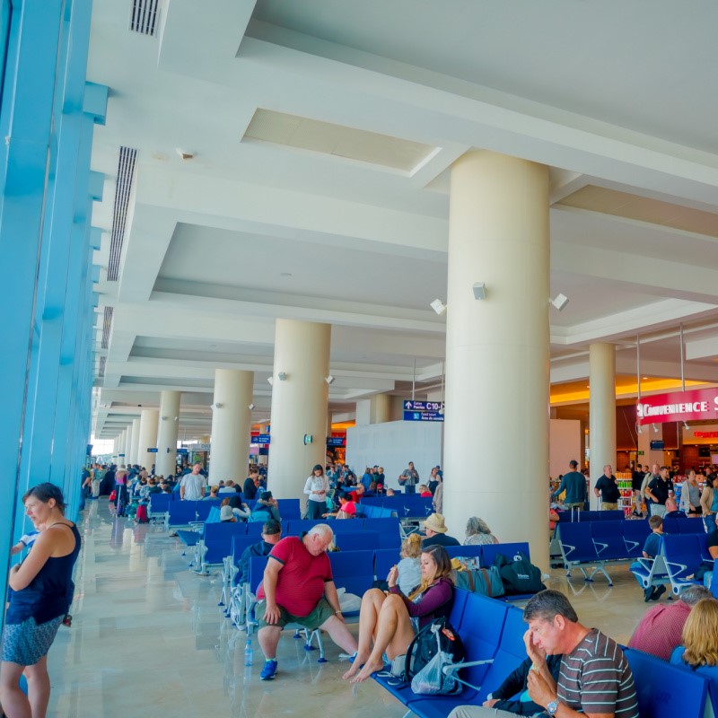 Viajeros que esperan vuelos en el aeropuerto de Cancún. Una mujer está en el fondo mirando por la ventana, mientras otras están sentadas y esperando.