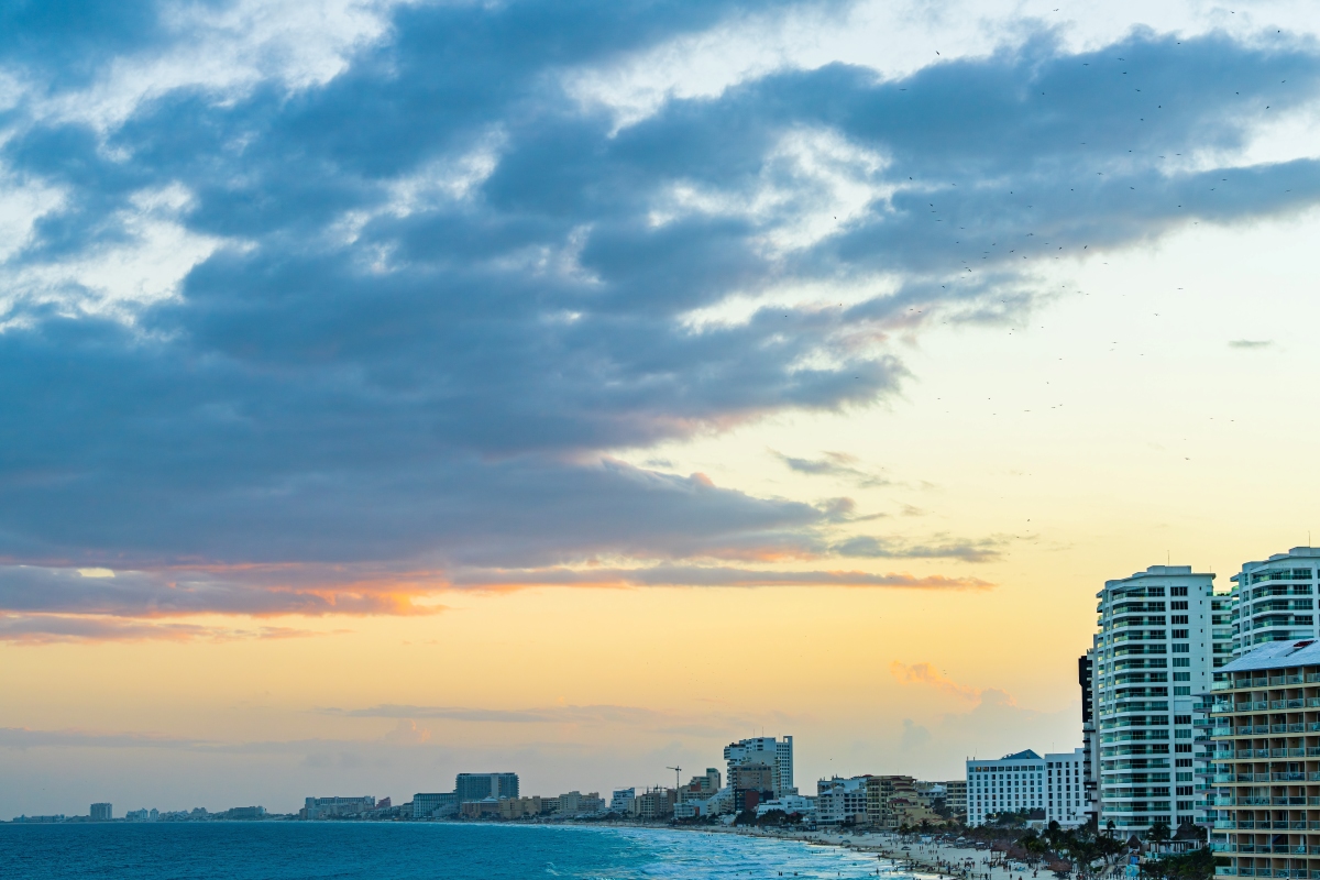 Una puesta de sol arroja un brillo cálido sobre la playa de Cancún, con nubes dispersas que reflejan la luz que se desvanece sobre la vibrante y bulliciosa orilla.
