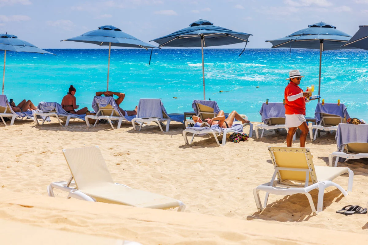 Playa caribeña con turistas relajándose en tumbonas bajo sombrillas azules mientras el camarero sirve bebidas contra las olas del mar turquesa. México. Cancún